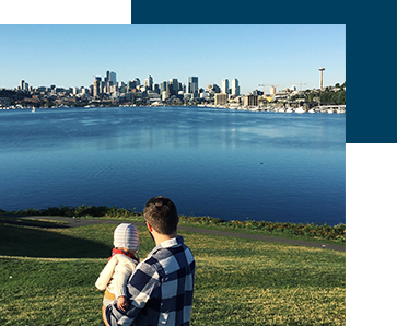 A person holding a small child is overlooking a large body of water with a city skyline in the background on a clear day. The scene includes grassy land in the foreground and multiple tall buildings in the distance.