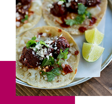 Tacos topped with meat, crumbled cheese, cilantro, and sauce, served on a metal plate with lime wedges on the side.