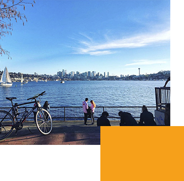 A scenic view of a waterfront with a city skyline in the background. A bicycle is parked by a railing, and people are sitting and standing nearby, enjoying the sunny day. A sailboat is visible on the water, and the sky is clear and blue.