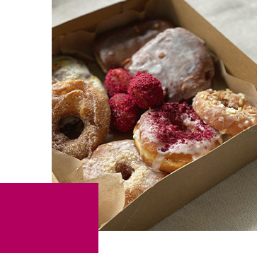 A box of assorted donuts, including glazed, sugar-coated, and ones topped with red sprinkles, sits on a light surface. Small pink cake balls are nestled among the donuts. The box is made of brown paper.
