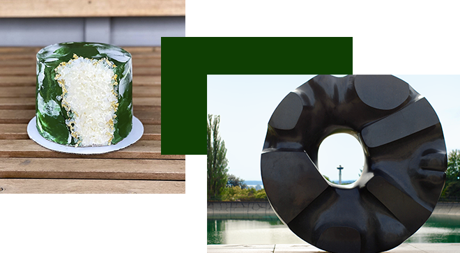 A green and white cake with a large, textured design sits on a wooden table. Beside it, a black abstract sculpture with a central hole is placed outdoors, with a blurred background of trees and the sky.