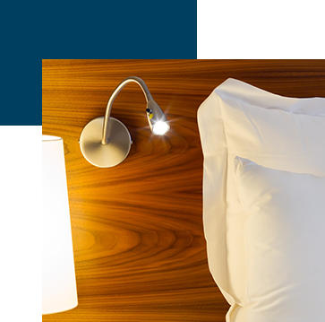 A modern hotel room scene features a wooden headboard with a mounted reading light turned on, illuminating white pillows and sheets. A portion of a lampshade is visible on the side.