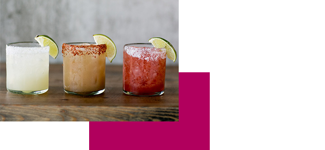 Three cocktails in glasses with salted rims sit on a wooden table. Each drink is garnished with a lime wedge. The drinks have different colors: clear, light brown with chili powder on the rim, and pink.