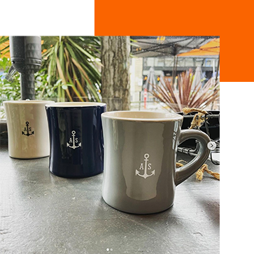Three mugs with anchor designs sit on a table. The mugs are white, navy blue, and gray. In the background, there are some plants and an outdoor setting with metal structures.