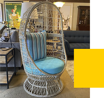 A wicker egg-shaped chair with a light blue cushion and striped pillow is displayed in a furniture store setting. A vintage-style poster and a blue sofa are in the background.