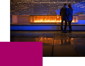 A couple stands closely together in front of a modern rectangular fire feature, surrounded by blue lighting. Their reflections are visible on the wet pavement.