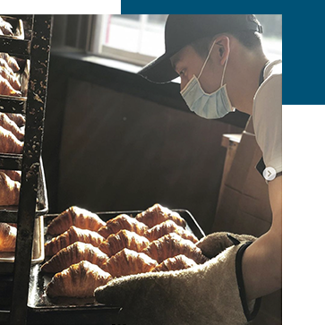 A person wearing a mask and cap, holding a tray of freshly baked croissants with oven mitts. The tray is being placed on a rack by a window, and the soft light highlights the golden-brown pastries.