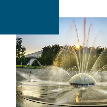 A dome-shaped fountain sprays water in various directions, surrounded by trees. The sun is setting behind the trees, casting a warm glow. In the background, a modern building with a slanted roof is partially visible.