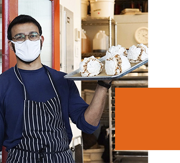 A person wearing glasses, a mask, and a striped apron holds a tray with four large desserts in a kitchen. An orange geometric shape is in the foreground.