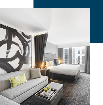 Stylish, modern hotel room with a king-sized bed, a gray sofa with a green cushion, and a matching ottoman. Large abstract art decorates the wall. Floor-to-ceiling windows let in ample light, offering views of skyscrapers outside.