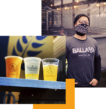 A person wearing a mask and a "Ballard Seattle, WA" shirt stands next to three cups of beer on a wooden ledge. The background features a building with string lights and a sign with "Reuben's Brews" logos on the cups.