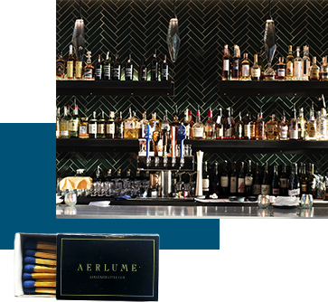 A sleek bar with dark herringbone tiles, lined with various liquor bottles on shelves and glasses on the counter. Below, a box of matches with "Aerlume" written on it is displayed.