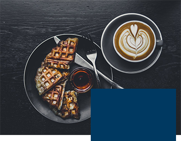 A plate of waffles with syrup and a fork on a dark table, accompanied by a cup of coffee with a heart-shaped latte art.