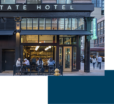 Street view of a hotel entrance with people sitting at a bar inside. The neon sign is visible, and pedestrians are walking by on the busy street.
