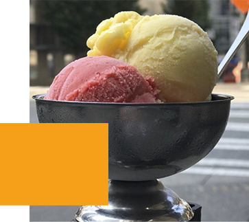 A metal bowl with two scoops of ice cream, one yellow and one pink, set outdoors on a sunny day. A spoon is in the yellow scoop. A blurred city street background is partially obscured by an orange geometric shape.