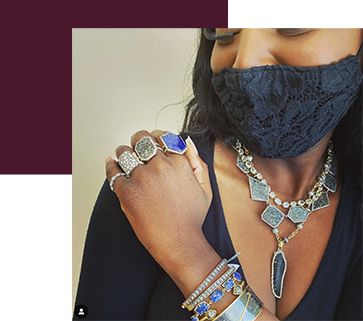 A person wearing a black lace mask showcases intricate jewelry, including a multi-layer necklace with stones, several large rings, and stacked bracelets. The image has a burgundy geometric border in the corner.