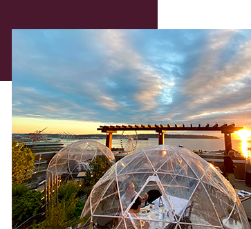 A scenic rooftop view at sunset with two transparent dining domes. The sky is a mix of blue and orange hues. Below, ships are visible in the water, and a wooden pergola frames the scene. People are sitting inside the domes.