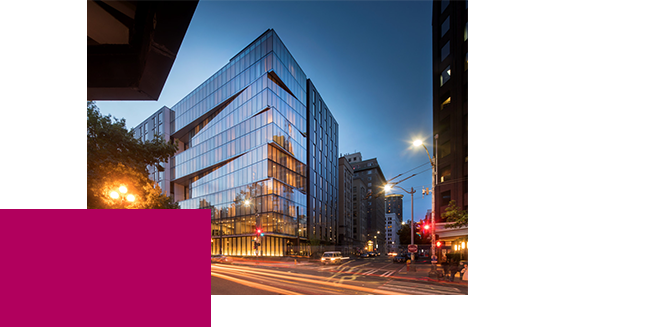 Modern glass office building at dusk with reflections of nearby buildings. Streetlights illuminate the intersection in front, and light trails from vehicles create dynamic lines. Adjacent structures and a purple geometric shape are also visible.